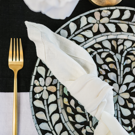 Detail of black and white mother of pearl inlay floral design on statement charger plate. White linen napkin knotted and set at place setting next to gold fork and 100% black linen table runner