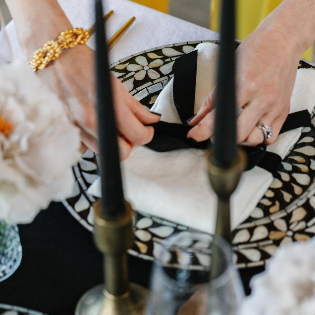 Laying Truffle Tablescapes' Magical Monochrome tablescape in a box with a classic 100% black linen table runner, black and white mother of pearl charger plates, antique brass candlesticks and black tapered dinner candles, and 100% linen white napkin and black velvet bows