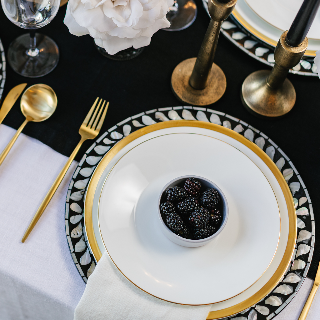 Black and white mother of pearl Vietnamese charger plate at place setting of monochrome tablescape. Gold trimmed plates and small bowl of blackberries next to gold cutlery and antique burnished brass candlesticks