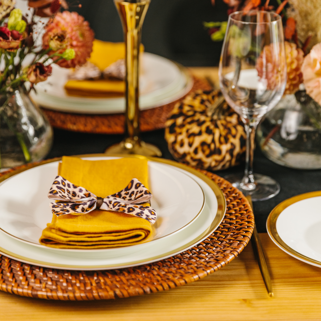 Autumnal place setting with brown rattan charger plate, ochre napkins, leopard print napkin bows and leopard pumpkin in background