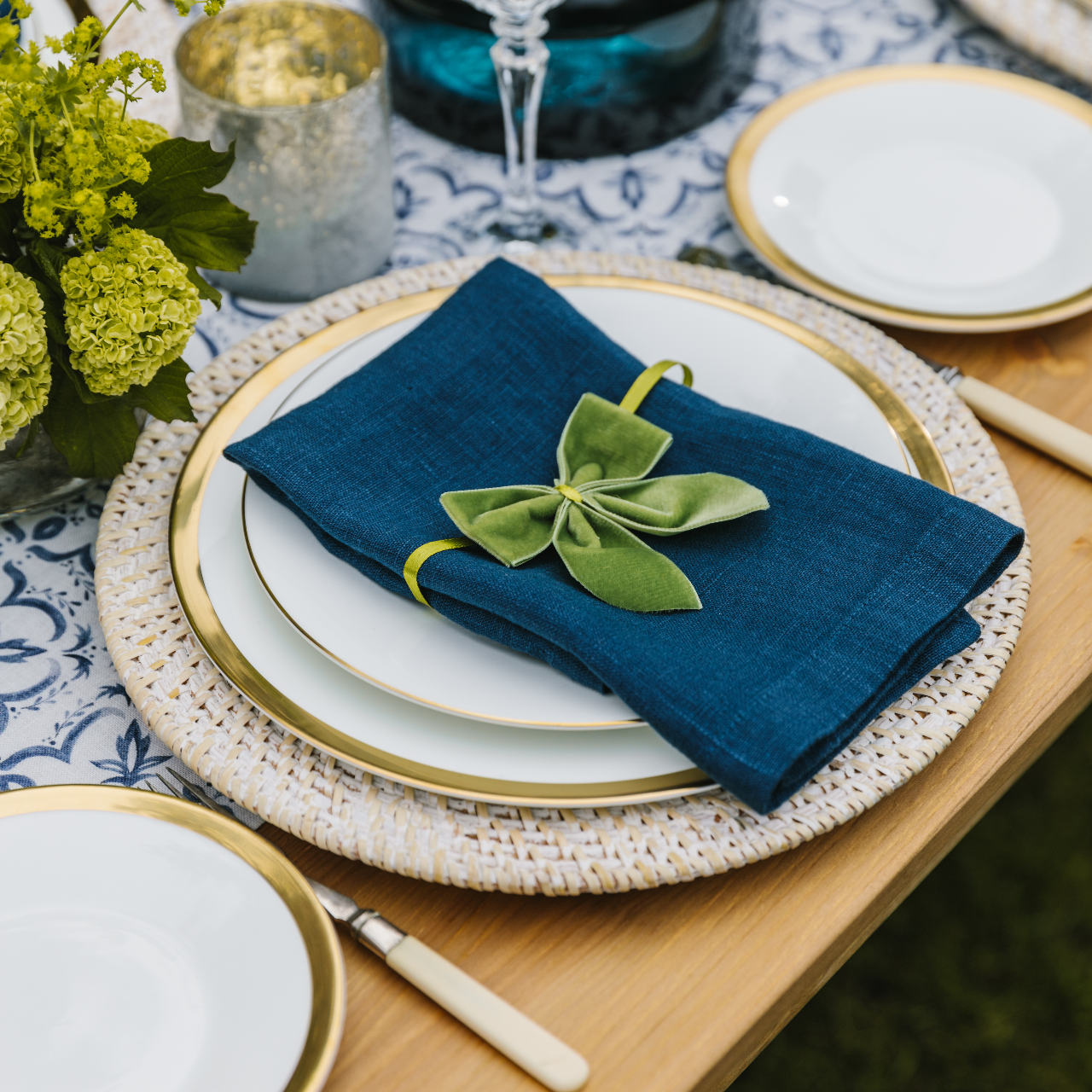 Navy linen napkin folded at a place setting and tied with a moss green velvet napkin bow. The place setting includes a white wash rattan charger plate, gold trimmed crockery and a blue and white mosaic linen table runner.