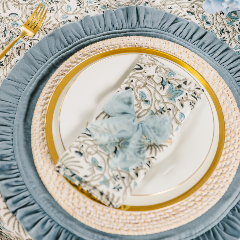 Bourbon blue velvet ruffled placemat place setting with white wash rattan charger plate, gold trim crockery, a folded Indian block print napkin and pale blue raw silk napkin bow.
