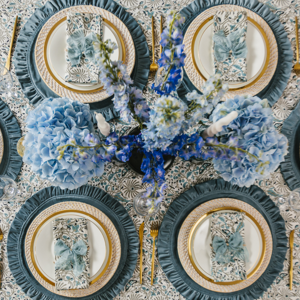 Top down view of Truffle's blue Twilight Collection including large blue flower central display and white and grey twist candles. The four place settings include bourbon blue velvet ruffled placemats, white wash rattan charger plates and Indian block print table linens.