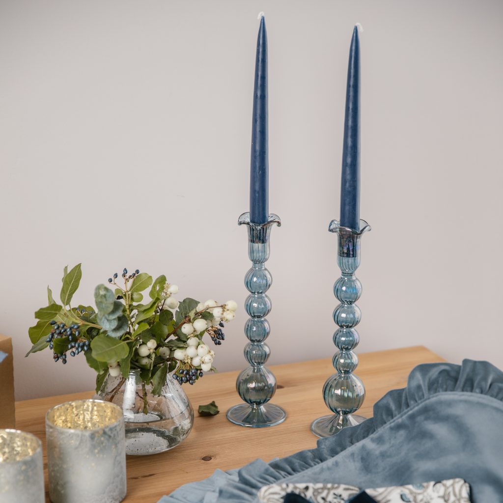 Two Egyptian hand blown candle holders set with two navy blue tapered dinner candles on a wooden table next to a bud vase filled with white and green flowers.