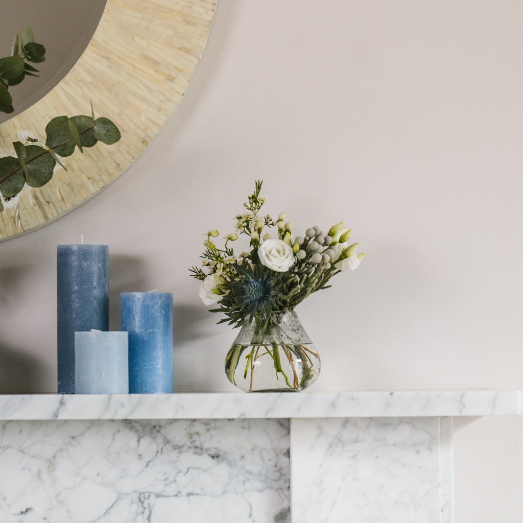 Distressed glass bud vase displaying blue and white flowers on white marble mantle next to cluster of blue toned pillar candles.