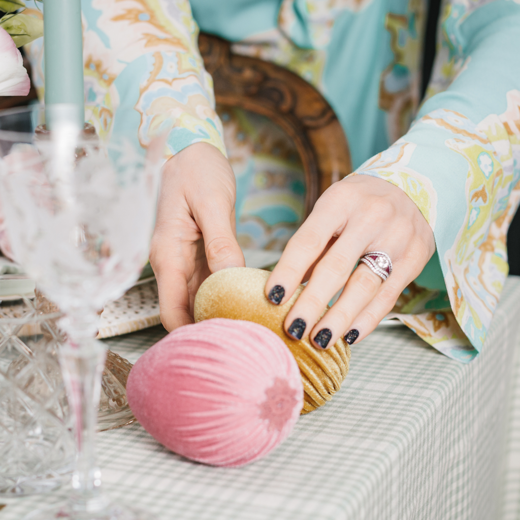 Hands placing a large pale gold velvet Easter egg next to a pink velvet Easter egg decoration on top of a duck egg gingham tablecloth and champagne gold glass candlestick