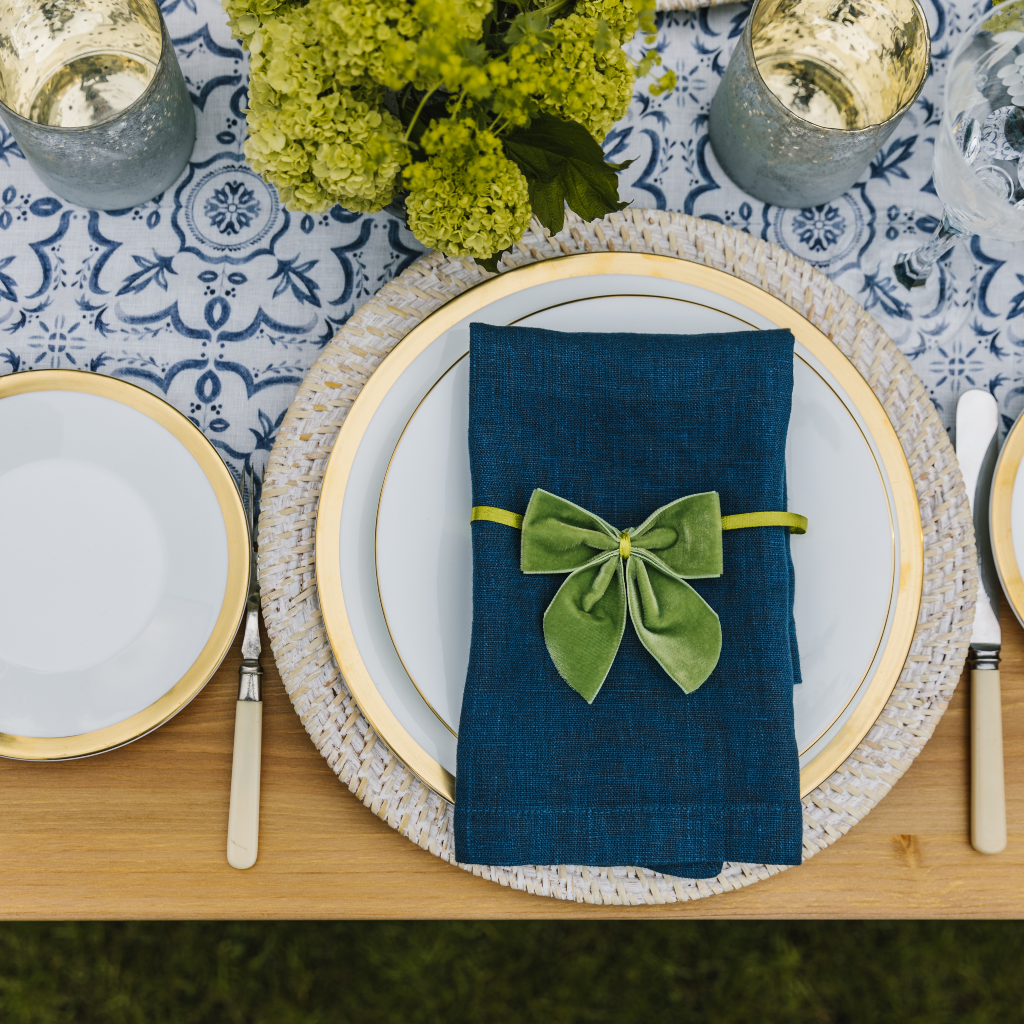 A statement blue and white mosaic linen table runner set on a table with one place setting including a white washed charger plate, white crockery with a gold trim, a navy linen napkin and moss green velvet napkin bow. Two golden tealight holders and a green posy of flowers are also displayed on top of the runner.