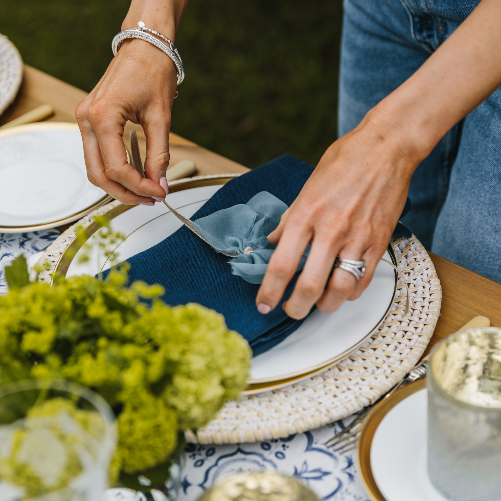 Female hands tying a vintage blue raw edge silk napkin bow round a 100% linen navy blue napkin on a place setting with white wash charger and white and gold plates.