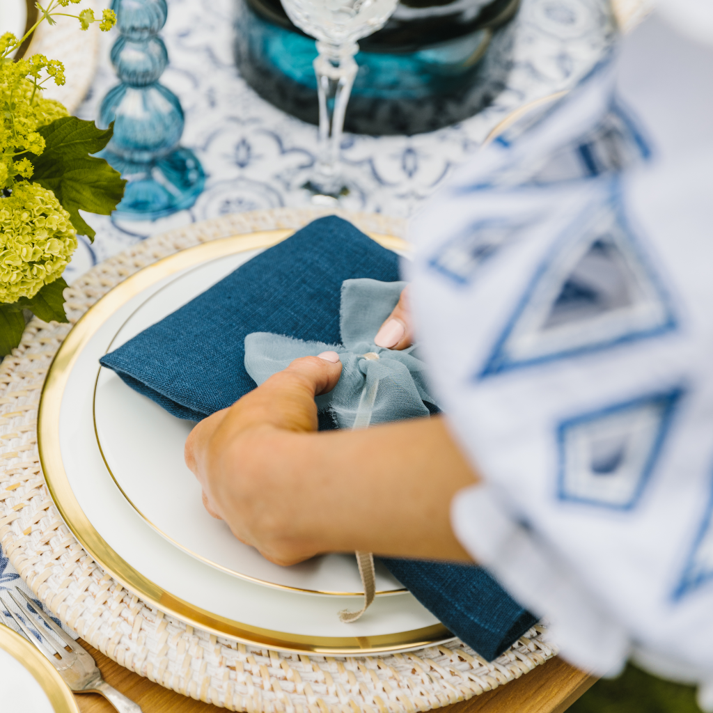 Hands laying a navy blue linen napkin at a place setting and adding a raw edge pale blue silk napkin bow. 