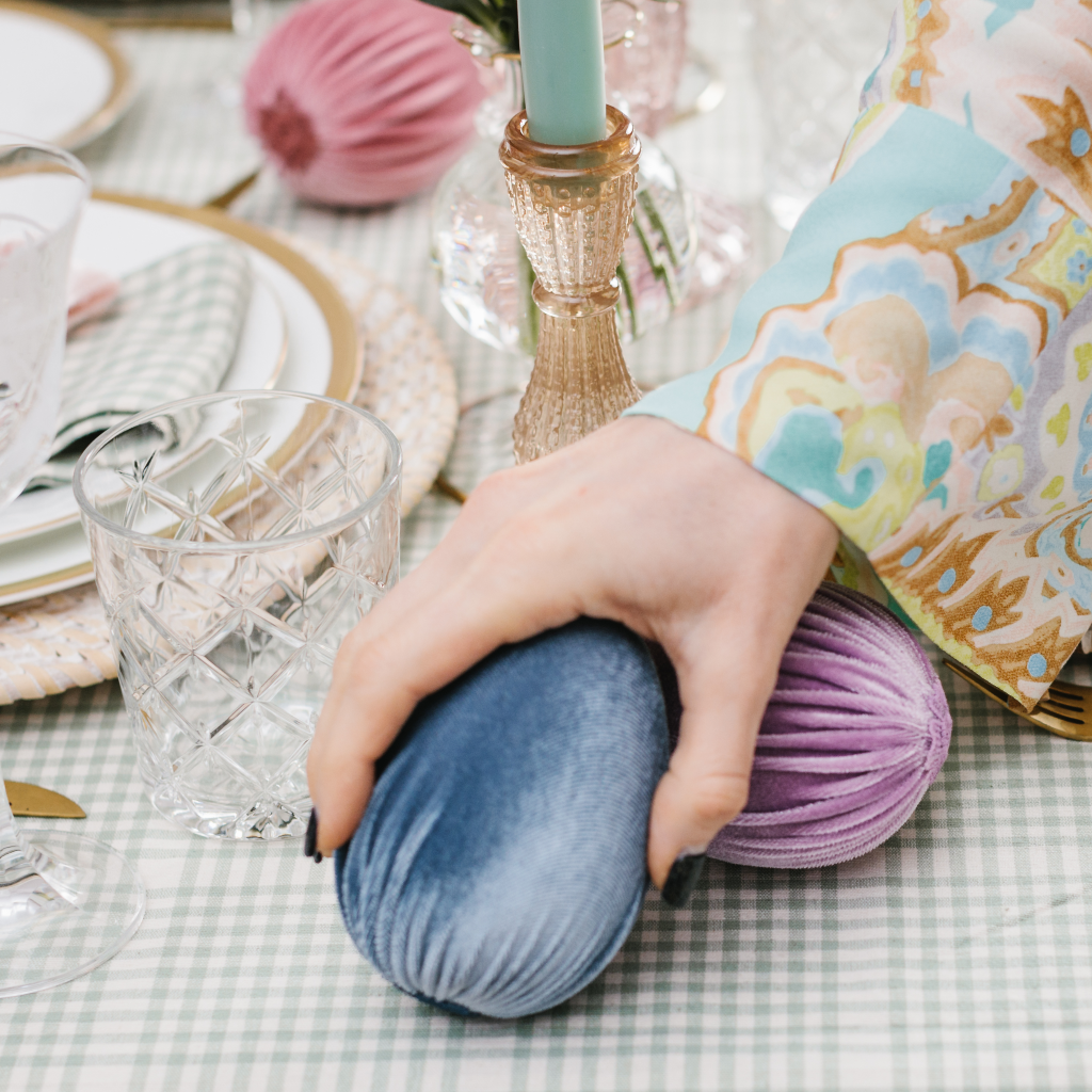 Hand holding a large slate blue handmade velvet Easter egg and placing it next to a cut glass tumbler on a blue and white gingham tablecloth