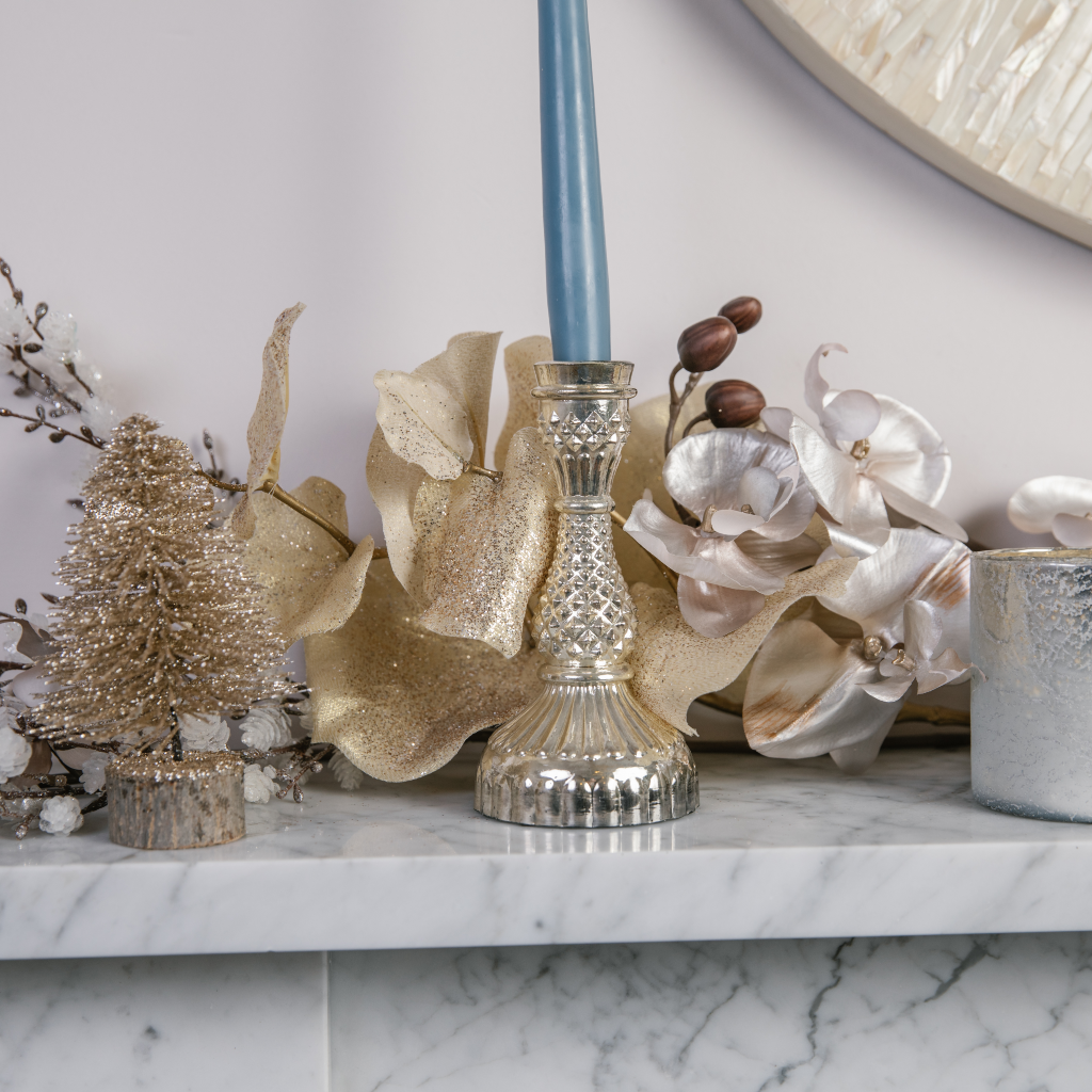 Ornate silver detailed candle holder with grey blue tapered candle next to gold glitter Christmas tree decoration on marble mantle