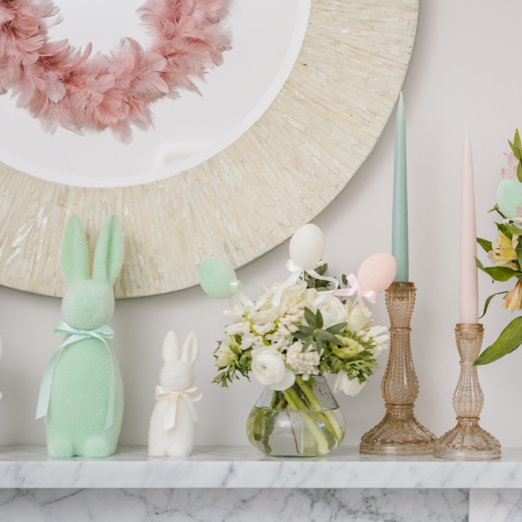 Clear glass bud vase containing a posy of cream flowers and flocked Easter egg decorations set next to soft gold glass candlesticks, pastel tapered candles and flocked Easter bunny decorations