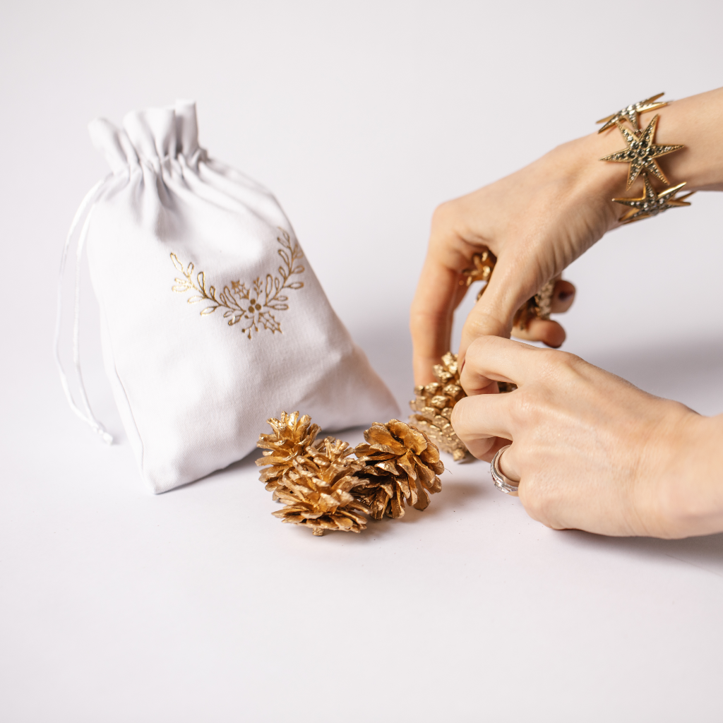 Hand arranging golden pine cones next to cotton pouch with gold holly wreath design