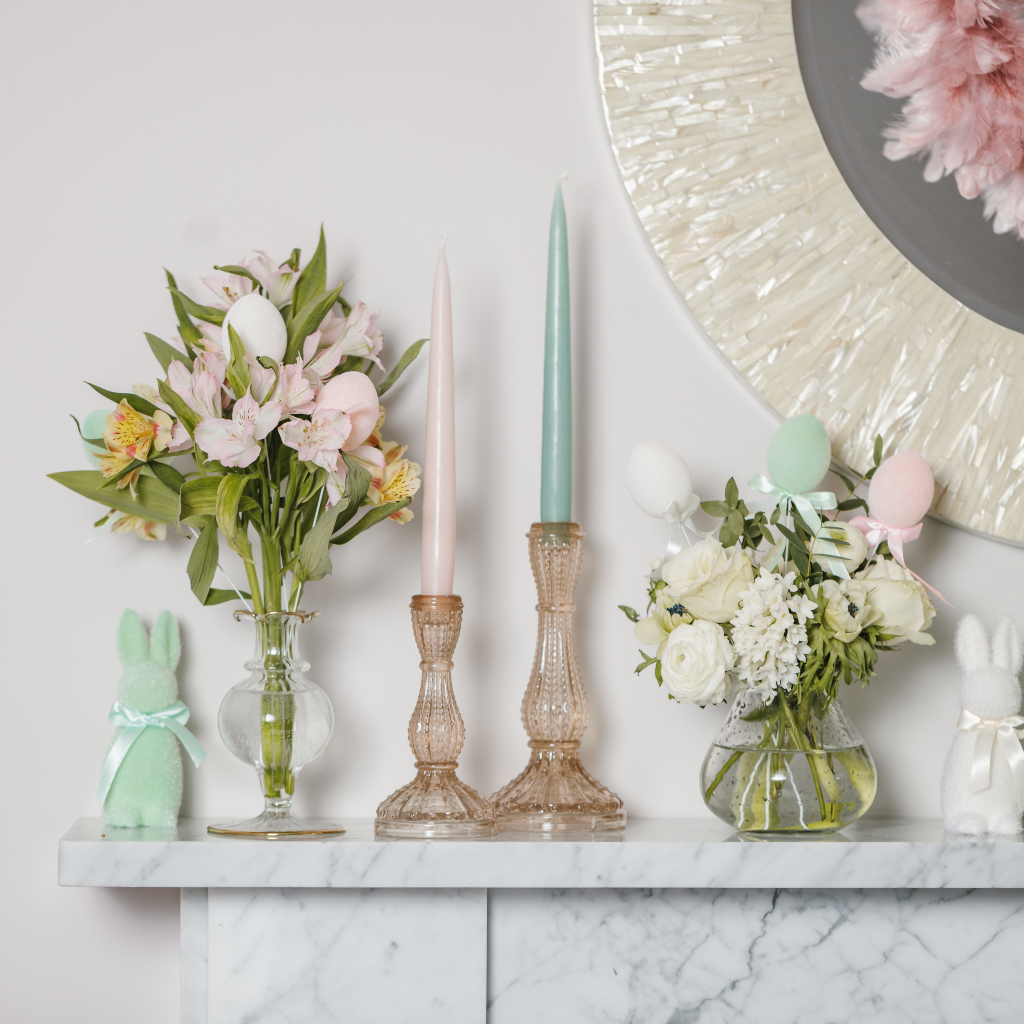 Clear glass bud vase containing a posy of cream flowers and flocked Easter egg decorations set next to soft gold glass candlesticks, pastel tapered candles and flocked Easter bunny decorations