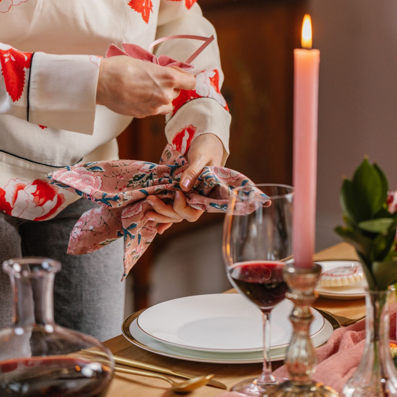 Hands tying a pink cotton napkin with peony flower pattern next to a pink dinner candle, burnished candle holder and carafe of red wine.