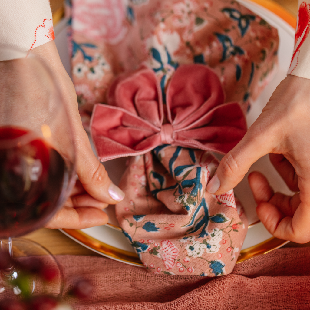 Hands placing a pink patterned cotton napkin on top of a plate next to a glass of red wine. A large pink velvet napkin bow clinches the napkin in at the centre forming a central pleat.
