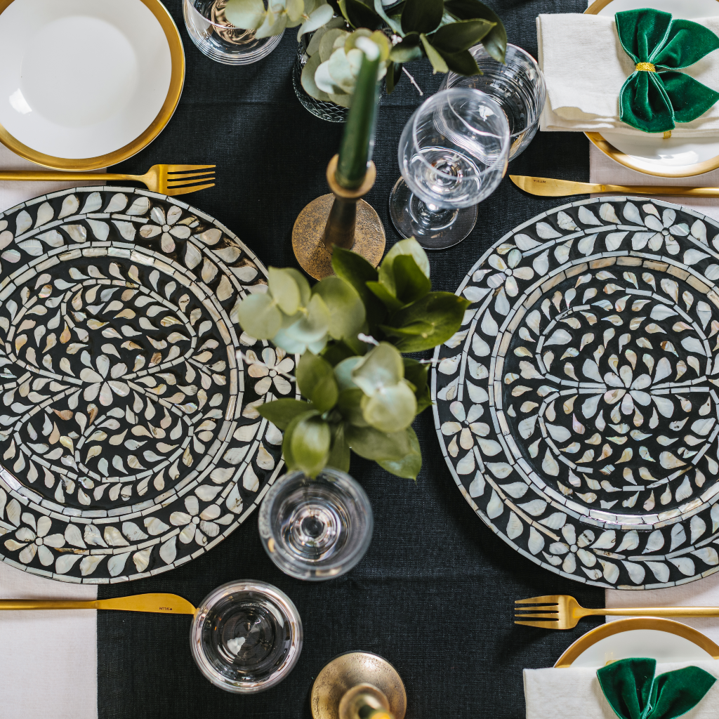 Top down flat lay image of black linen table runner, statement black and white mother of pearl inlay charge plates, brass candlesticks, forest green tapered candles, luxe green and gold velvet napkin bows and festive greenery in bud vases