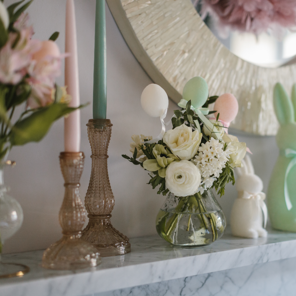 Champagne gold cut glass candle holders with powder pink and sage tapered candles next to a posy of cream flowers and flocked Easter eggs with ribbon ties