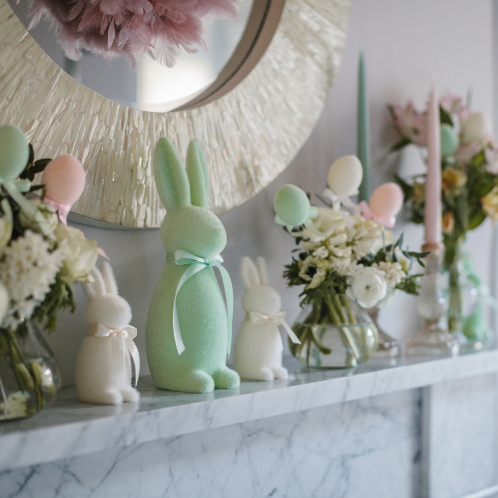Mint green large Easter bunny sitting between two small cream flocked Easter bunnies on a white marble mantle. Clear glass bud vases with posies of flowers and flocked Easter egg decorations are displayed next to champagne gold candle holders with powder pink and sage green tapered candles.