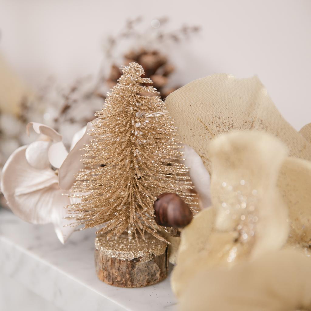 Champagne gold glitter tree decoration with wooden base placed next to metallic festive foliage