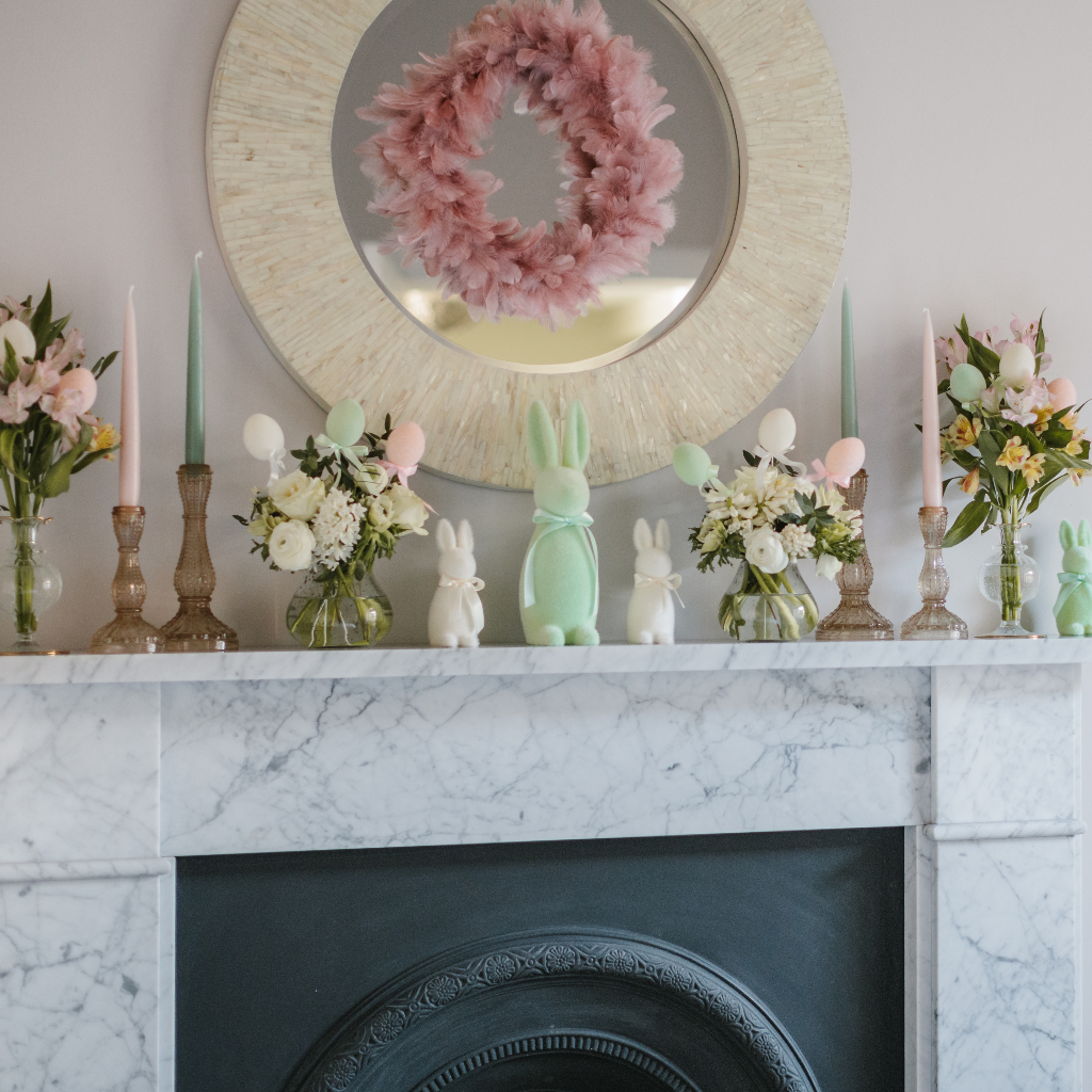 Easter mantlescape featuring a large mint green Easter bunny, small cream flocked Easter bunnies. posies of spring flowers displayed in hand blown bud vases and champagne gold glass candle holders with tapered candles