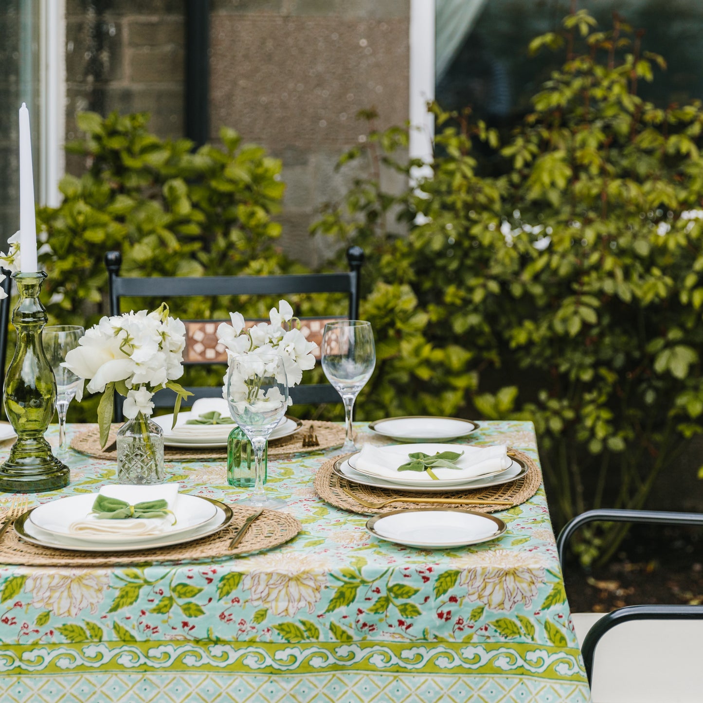 View of Mint & Moss tablecloth set outdoors with seagrass placemats, clear cut glass bud bottles, pale green bud bottles and large green glass candlesticks
