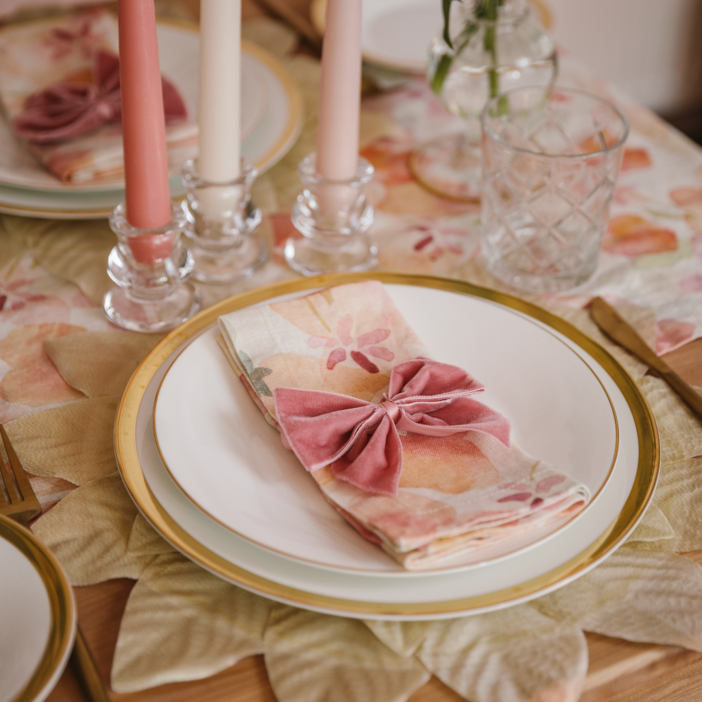 A champagne gold leaf placemat set with gold trimmed plates and a pink, green and peach floral linen napkin and matching table runner. Mini tulip clear glass candle holders display a trio of vintage rose, off white and powder pink tapered candles set next to a hand blown gold trimmed bud vase