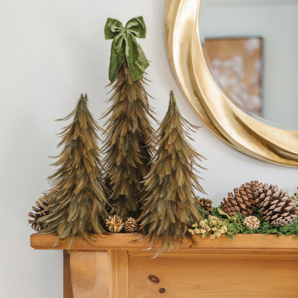 Large feather Christmas tree decoration topped with velvet bow flanked by two medium feather Christmas trees on mantle with giant pine cones and moss