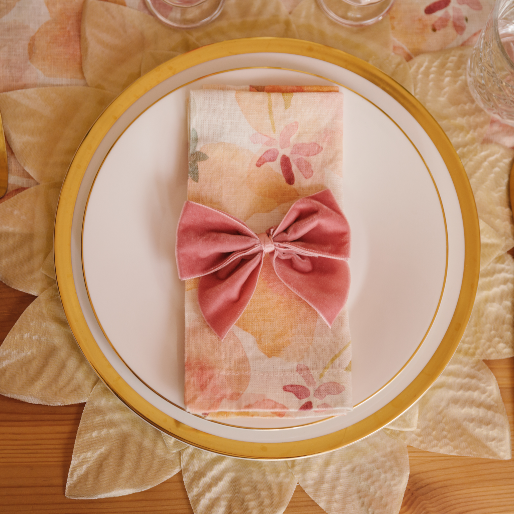 Top down image of table place setting featuring champagne gold leaf placemats, gold trimmed plates, pink, peach and cream watercolour patterned linen napkins and a pink velvet napkin bow