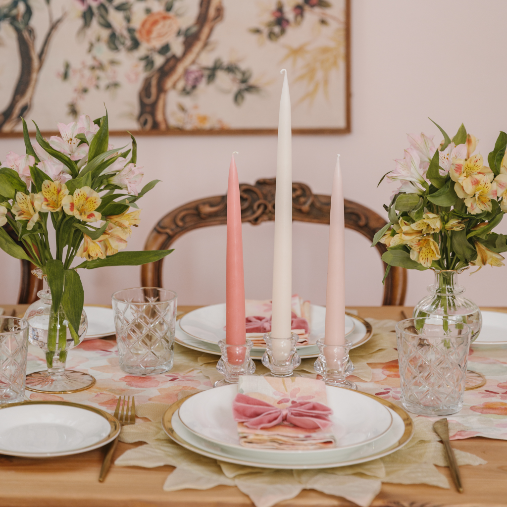 The Peaches and Cream tapered dinner candle set including a vintage rose pink, an off white and powder pink set of tapered candles. The candles are displayed in small glass tulip candle holders next to clear bud vases with a gold trim. The table is also set for two with peach, pink and cream linen napkins and table runner and luxury pink velvet napkin bows