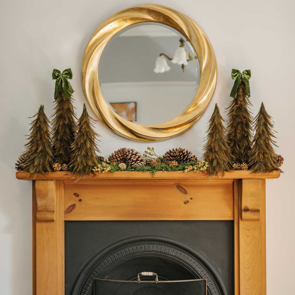 Traditional wooden mantlepiece with feather Christmas tree display alongside golden pine cones and giant pine cones under large gold mirror