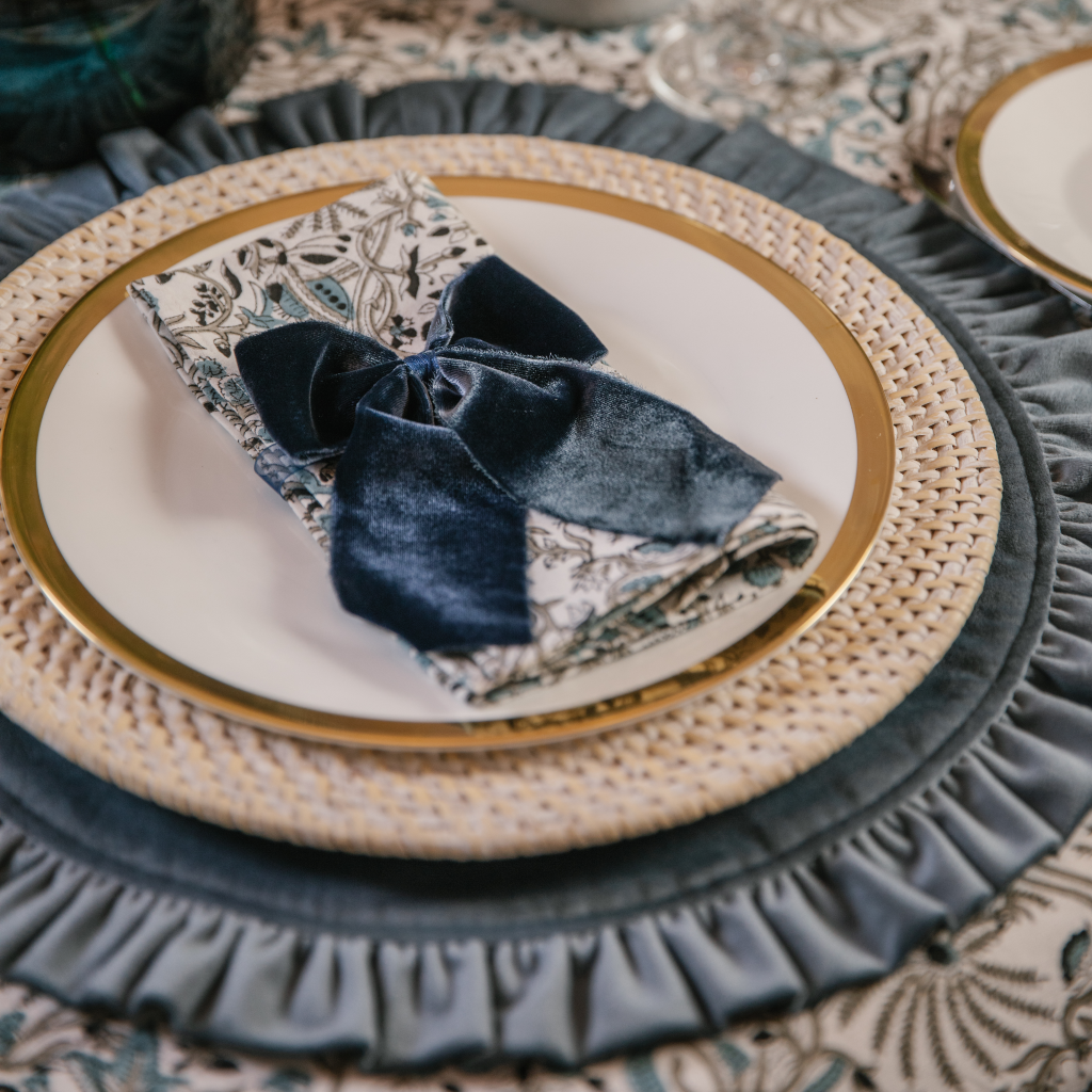 Blue tablescape place setting with ruffled placemat, white wash rattan charger plate, Indian block printed napkins and tablecloth and  navy blue velvet napkin bows.