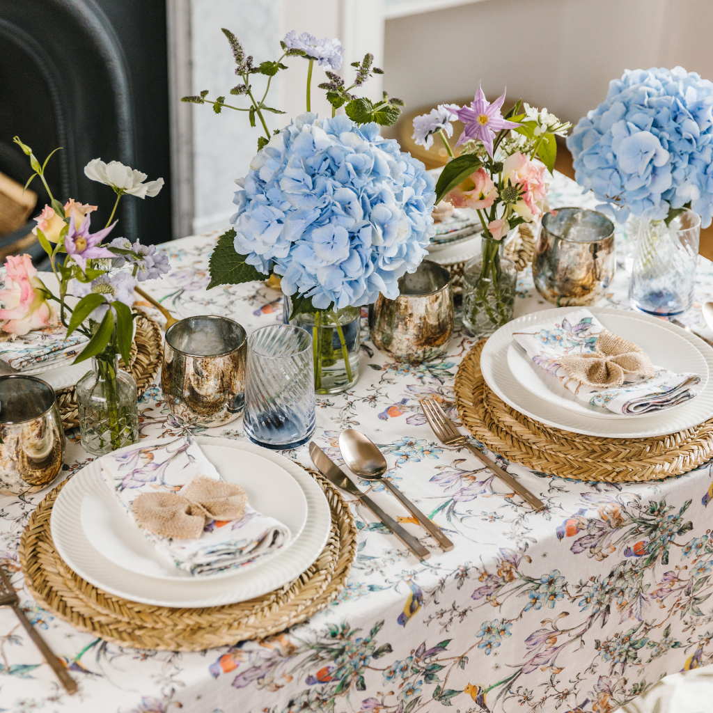 Wildflower Garden tablescape from a side-on view. 100% linen tablecloth with birds and wildflower pattern, seagrass placemats, white crockery, wildflower napkins and hessian bows. All finished with giant blue hydrangea and copper burnished candle holders.