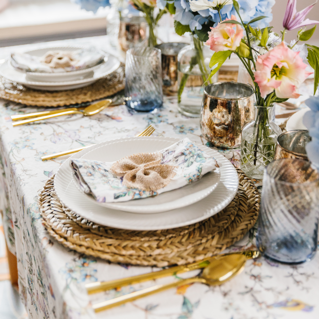 Wildflower Garden tablescape from a side-on view. 100% linen tablecloth with birds and wildflower pattern, seagrass placemats, white crockery, wildflower napkins and hessian bows. All finished with giant blue hydrangea and copper burnished candle holders.