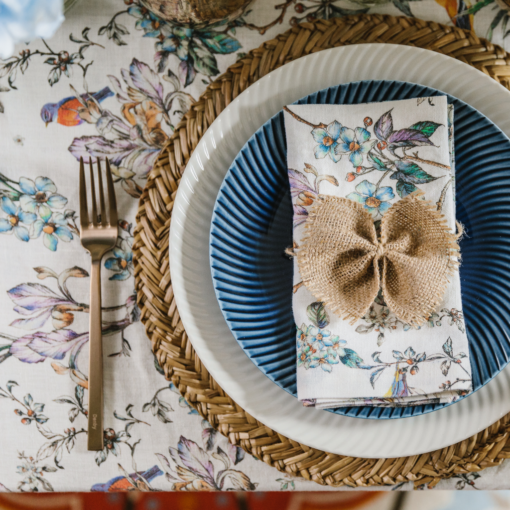 Top down view of Wildflower Collection place setting including patterned linen napkins and hessian napkin bow, Denby porcelain crockery, seagrass placemat and white tablecloth with flower pattern.