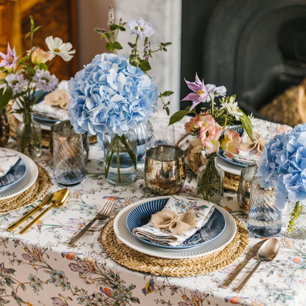 Wildflower Garden tablescape from a side-on view. 100% linen tablecloth with birds and wildflower pattern, seagrass placemats, white crockery, wildflower napkins and hessian bows. All finished with giant blue hydrangea and copper burnished candle holders.