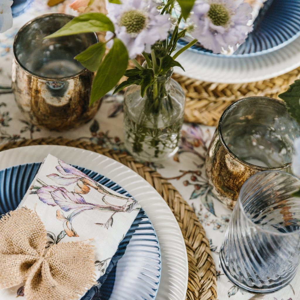 Close-up view of copper burnished tealight holders. cut glass bud vase and wildflower place setting. A large hessian napkin bow is tied round a patterned linen napkin set on blue and white crockery and a seagrass placemat.