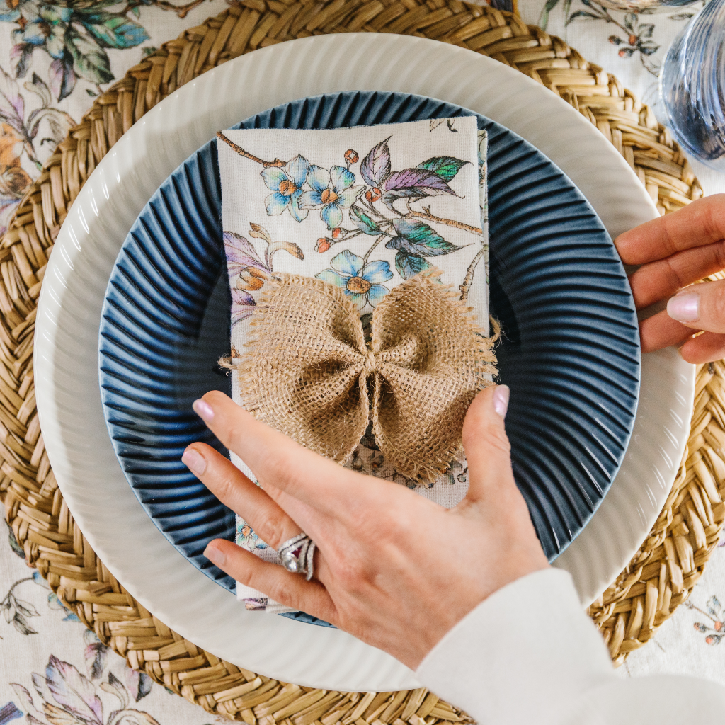 Top down view of hand tying hessian bow napkin ring round wildflower and bird printed linen napkin. The place setting including Denby Porcelain crockery in navy and white with an arc pattern and natural seagrass placemats.