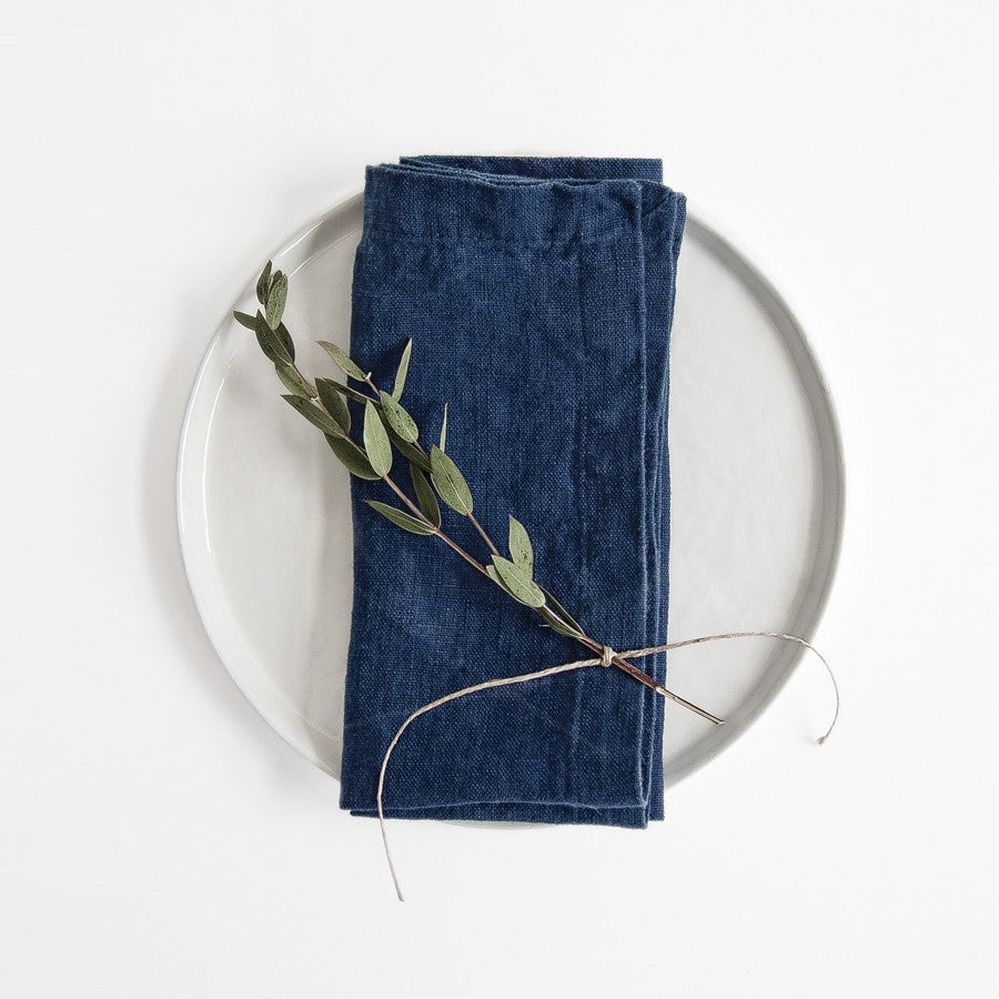 A dark navy linen napkin folded on a white dinner plate with one sprig of greenery placed on top.